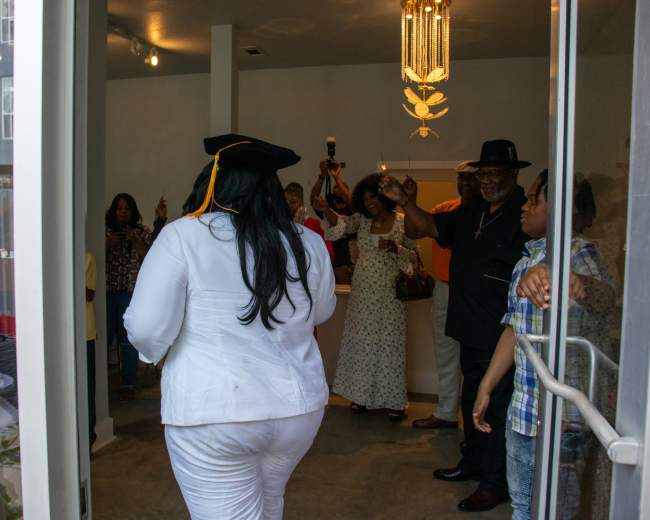 A woman in a white outfit and graduation cap walks through a doorway while a group of people celebrate inside.