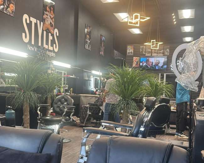 The image shows a modern barbershop interior with black styling chairs, tropical plants, and a large television displaying news.