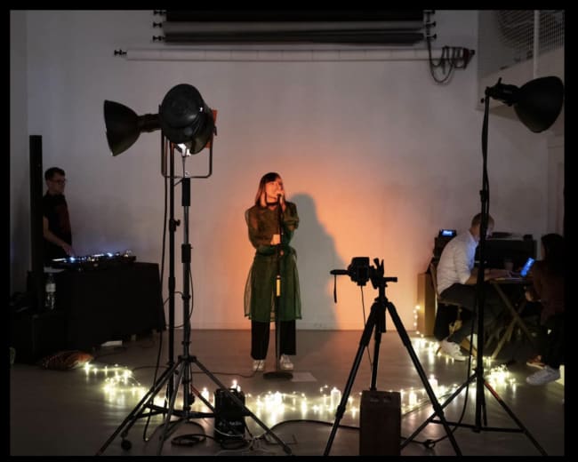 A woman stands on a stage with a microphone, surrounded by lights and camera equipment while a DJ performs in the background.