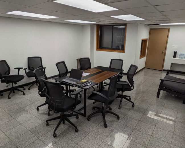 A conference room features a large rectangular table surrounded by several black ergonomic chairs on a gray tiled floor.