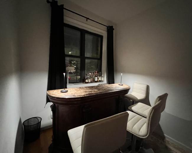 A small home bar area features a wooden bar top with two white stools, illuminated by two lamps, and a window visible in the background.