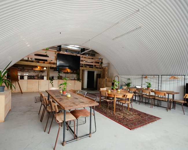 The image shows an industrial-style interior of a spacious venue with a curved metal ceiling, wooden furniture, and plants arranged on tables.