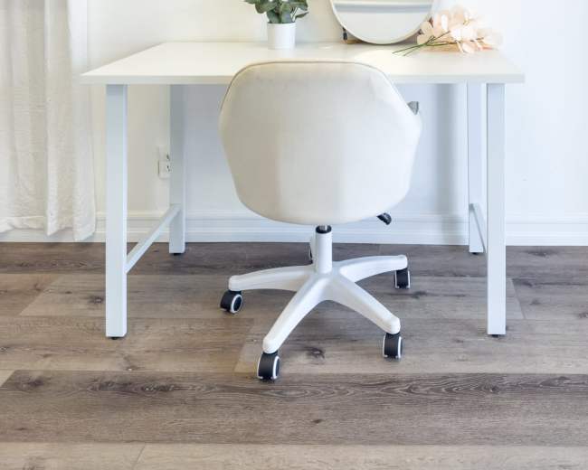 A white desk with a rolling chair is positioned in front of a circular mirror, accompanied by a small potted plant and a decorative flower on the desk.