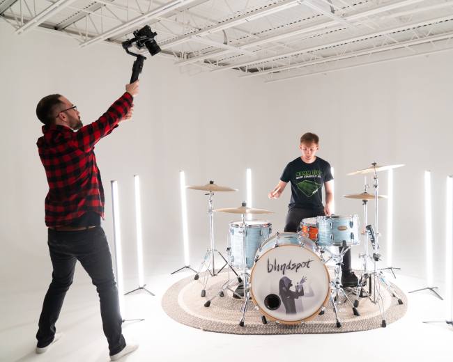 A drummer performs in a studio surrounded by drum equipment and illuminated by vertical light fixtures, while a man holds a camera on a stabilizer.