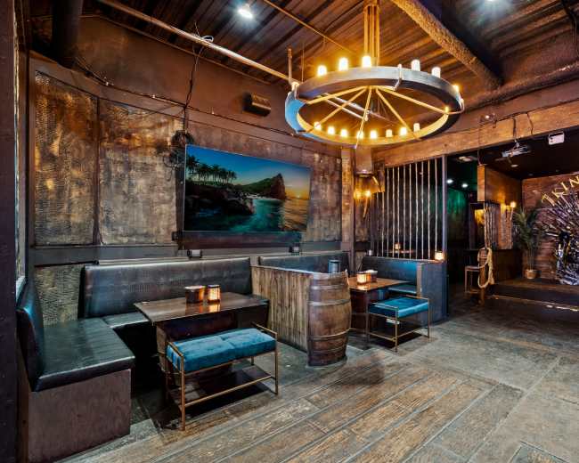 A dimly lit restaurant interior featuring wooden tables and benches, a large chandelier, and tropical-themed decor on the walls.