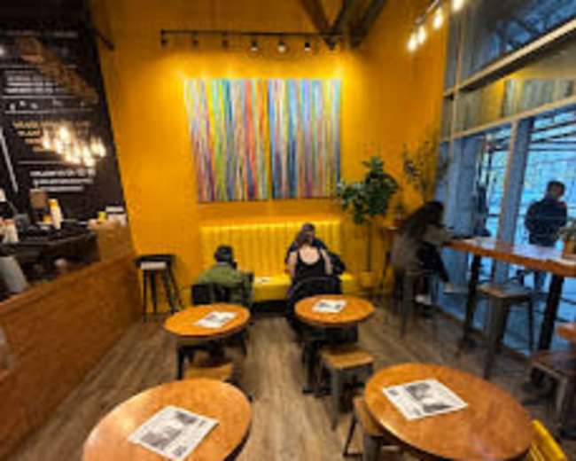 The image shows a cozy café interior with wooden tables and stools, yellow walls, and colorful artwork hanging above a seating area.