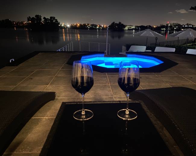 Two glasses of red wine sit on a table beside a hot tub with blue lighting, overlooking a calm body of water at night.
