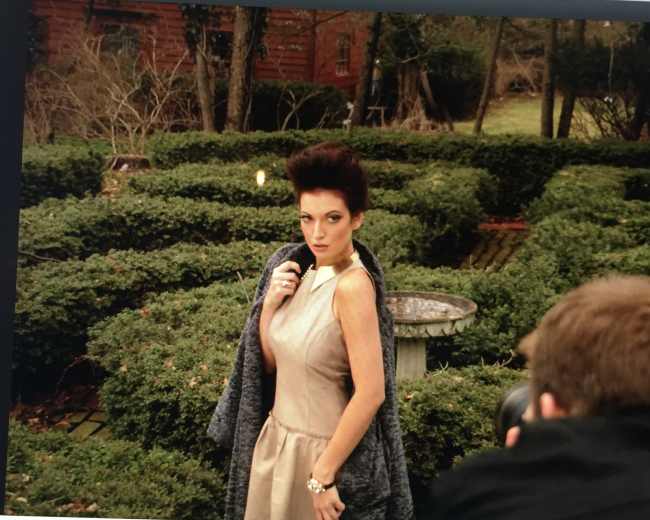 A model poses in a garden with neatly trimmed hedges while a photographer captures her image.