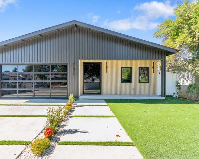 A modern house features a metal facade with a large glass garage door, surrounded by a landscaped yard and pathway.