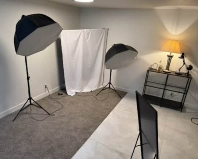 The image shows a photography setup in a room, featuring two softbox lights, a white backdrop, and a lamp next to a small table.