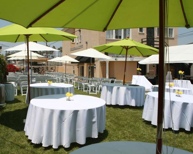 A setup of outdoor tables with white tablecloths and green umbrellas in a grassy area, surrounded by buildings and potted plants.