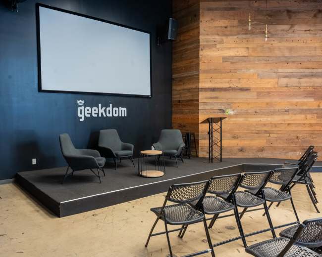 The image shows a small stage with three chairs, a table, and a podium, set against a wooden wall in a room labeled "geekdom."