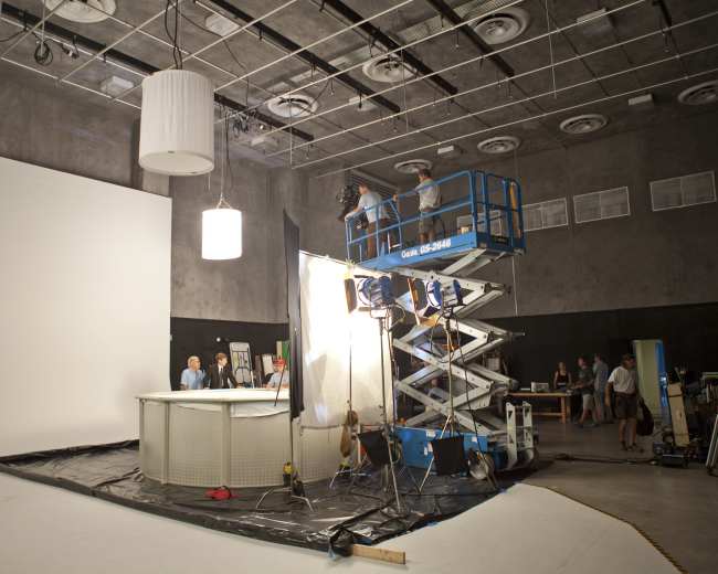 A film crew sets up lighting and equipment in a studio featuring a large white backdrop and a raised platform.