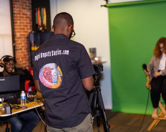 A video production setup features a person in a black shirt with a logo directing a seated individual in front of a green screen, while another person operates equipment nearby.