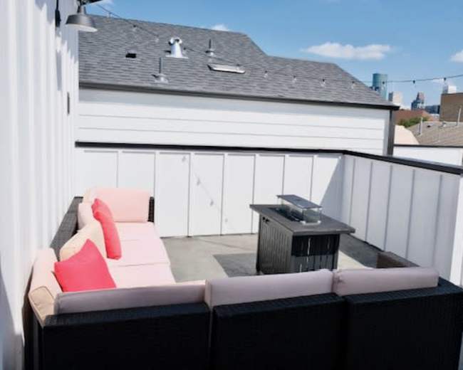A rooftop patio features a sectional sofa with pink cushions and a black fire pit surrounded by a white railing and views of the city skyline.
