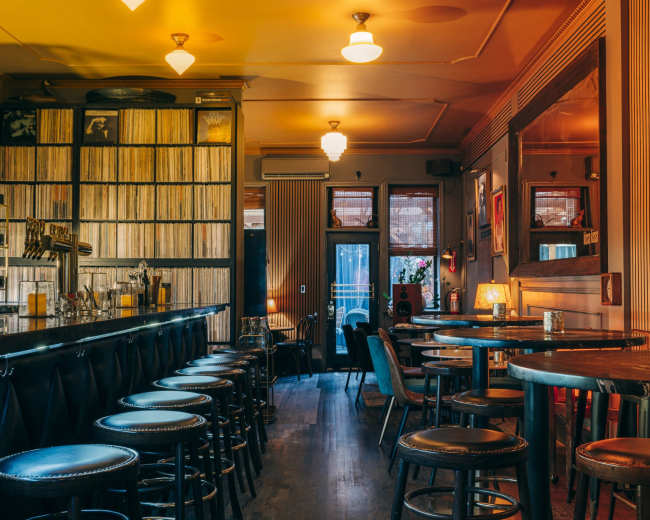 The image shows a dimly lit bar interior with a long bar counter, several round tables, and walls lined with vinyl records.