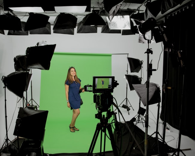 A woman stands on a green screen set surrounded by studio lights and camera equipment.