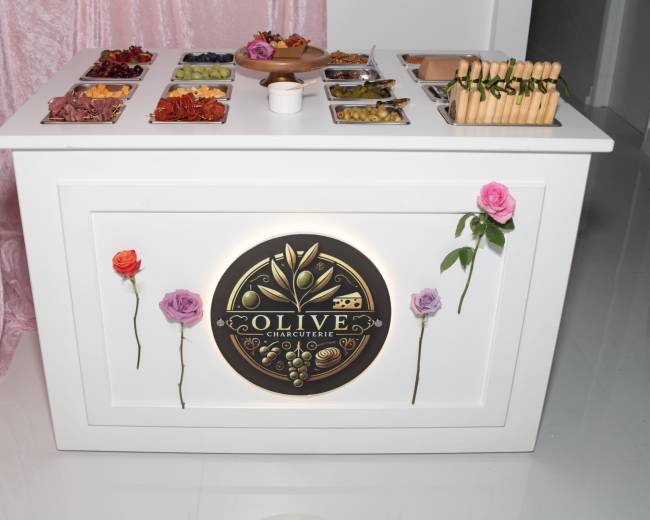 A white charcuterie table is adorned with a variety of food items in trays, featuring a decorative logo and fresh flowers on the front.