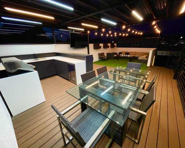 The image shows a modern rooftop patio with a dining table, outdoor kitchen, and lounge area, all illuminated by string lights at night.