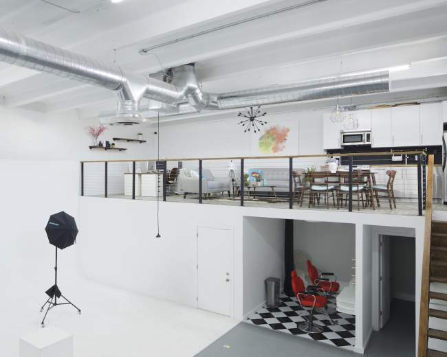 The image shows a spacious, modern studio with a loft area, featuring a seating area and dining space, and a lower level with black and white checkered flooring and two red chairs.