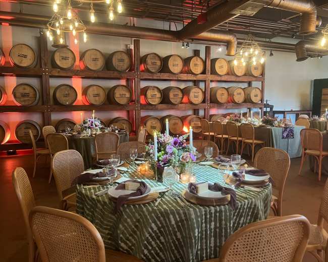 The scene features a dining area set with round tables adorned with floral centerpieces, surrounded by wine barrels against a softly lit wall.