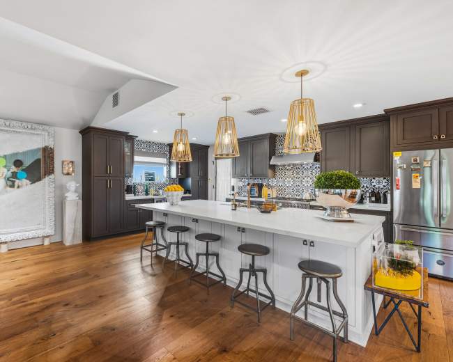 The kitchen features a large island with bar stools, modern cabinetry, and a decorative painting on the wall, all illuminated by stylish pendant lights.