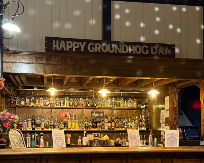 The image shows a wooden bar with a sign reading "Happy Groundhog Day" above shelves lined with various bottles of liquor, and empty bar stools in front.