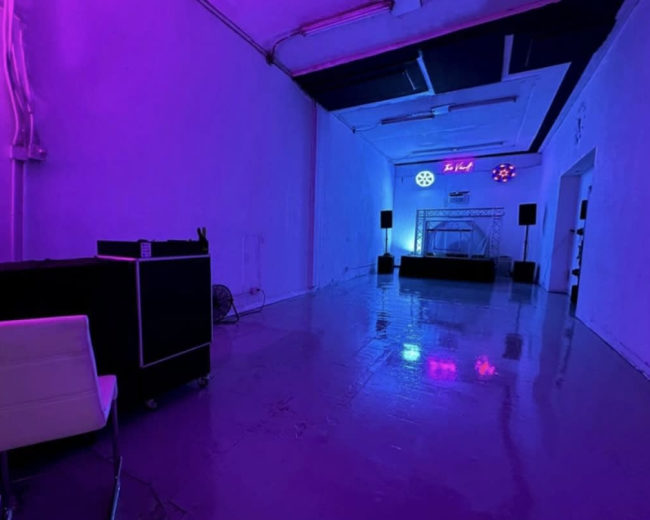 The image shows a spacious, dimly lit room with purple lighting, featuring a small stage at the far end and audio equipment along the walls.