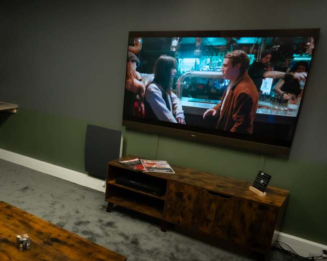 A media room is set up with two computer monitors, a white desk chair, a wooden coffee table, and a large television displaying a scene from a movie.