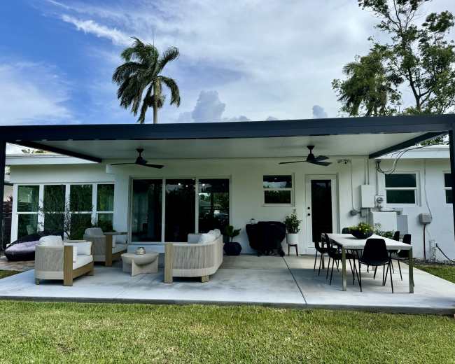 The image shows a covered outdoor patio area with a seating arrangement and dining table, surrounded by grass and palm trees.