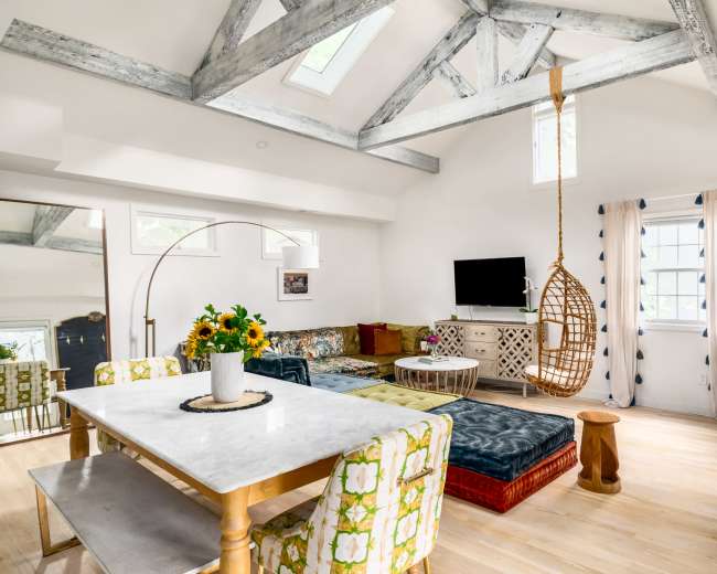 The image shows a bright, modern living space with exposed wooden beams, a hanging chair, and a dining table surrounded by decorative seating.