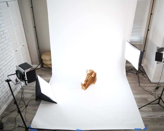 The image shows a photography setup with bright lights and a blank backdrop, featuring a wooden object positioned at the center.