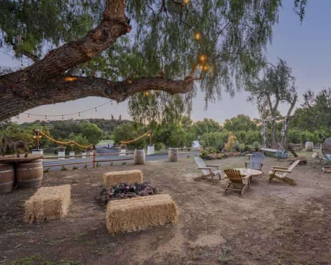 A fire pit is surrounded by wooden chairs and straw bales under a tree with string lights in an outdoor setting.