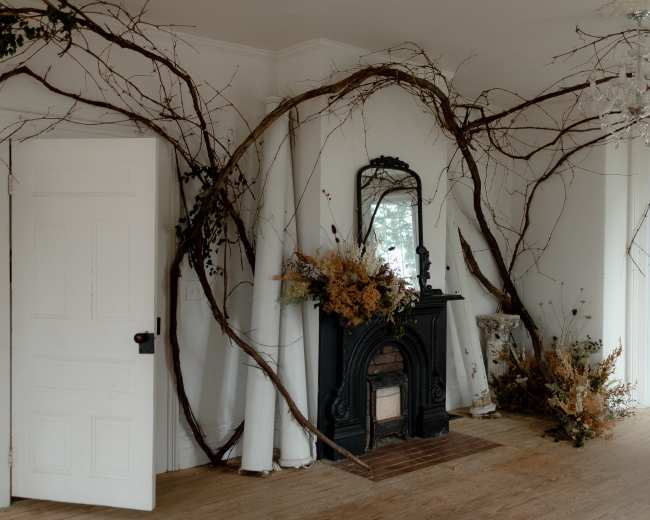 The room features a fireplace surrounded by large, twisted vines and dried floral arrangements, with a mirror reflecting the space.