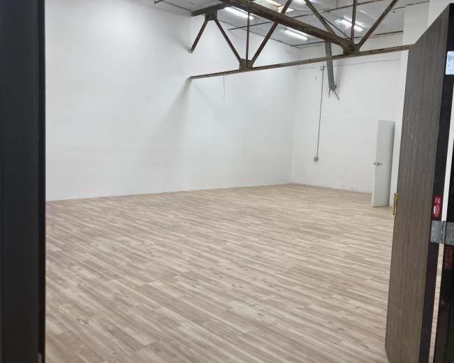 The image shows an empty room with wooden flooring and white walls, viewed from an open doorway.