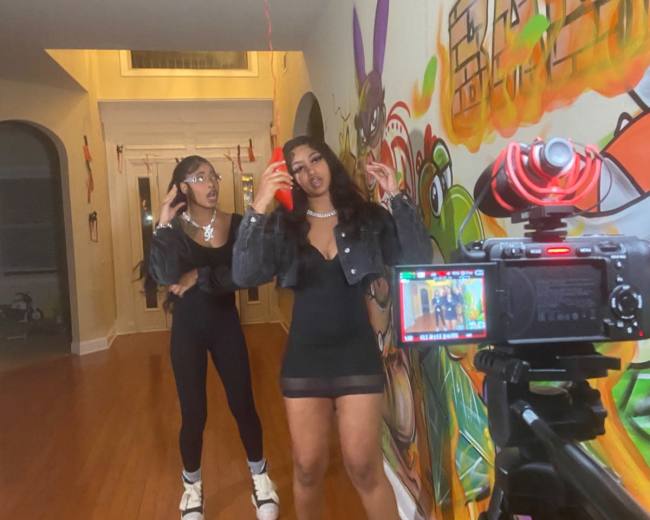 Two young women pose for a photo in a hallway adorned with colorful graffiti, while a camera on a tripod captures the scene.