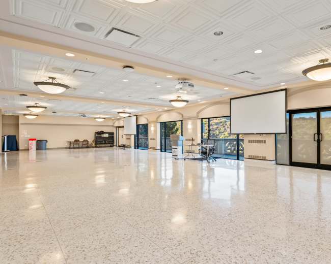 The image depicts a spacious, empty indoor hall with large windows, a podium, and two projection screens.