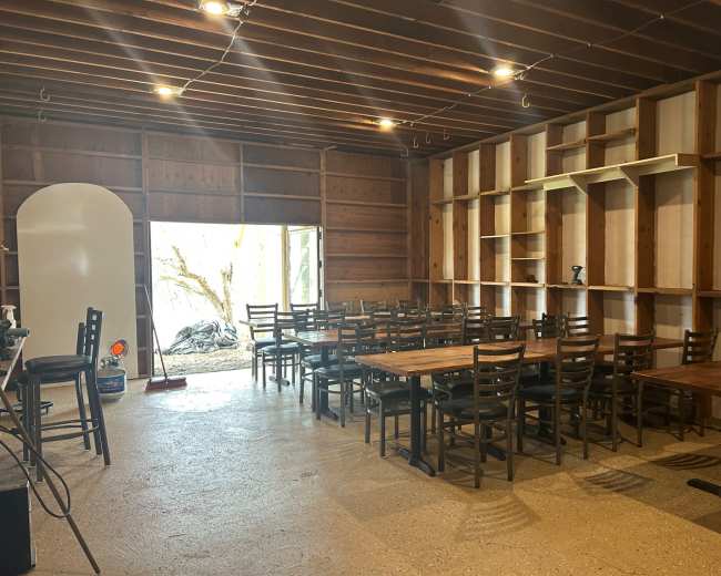 The image shows a spacious room with wooden walls, featuring rows of tables and chairs facing a large opening that leads to an outdoor area.