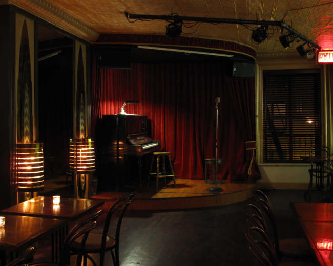 The image depicts a dimly lit performance stage in a small venue, featuring a red curtain, a vintage piano, and a microphone on a stand.