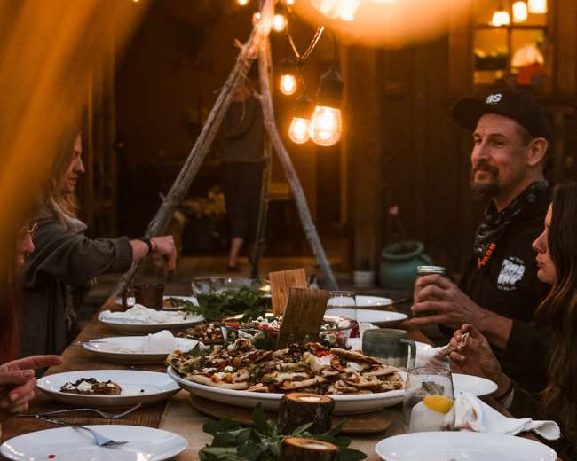 A long wooden table is set for a gathering with plates of food and decorative elements, surrounded by people in a warmly lit outdoor setting.