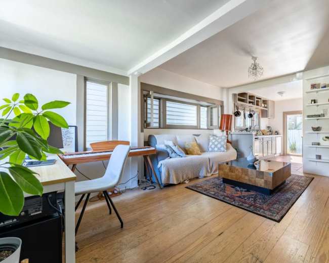 The image shows a bright living room with a sofa, coffee table, and a dining area featuring a bookshelf and kitchen in the background.