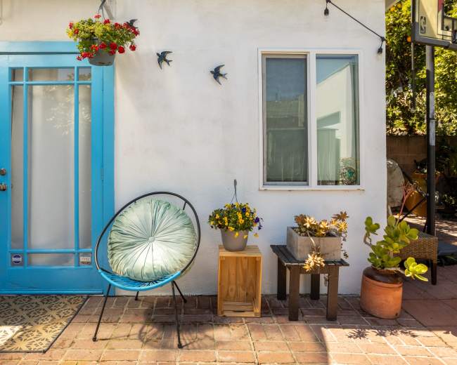 The image shows a patio area featuring a blue door, a circular chair, potted plants, and decorative birds on a white wall.