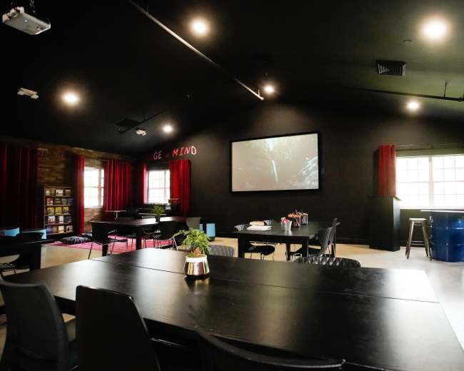 The image shows a spacious meeting room with a large screen, black walls, red curtains, and various seating arrangements, including tables and bar stools.