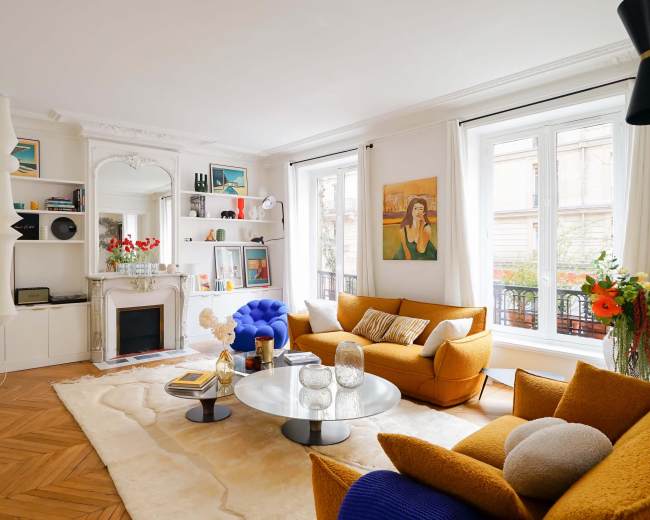 The image shows a bright living room featuring a yellow sofa, a blue armchair, a round coffee table, and large windows with views of the outdoors.