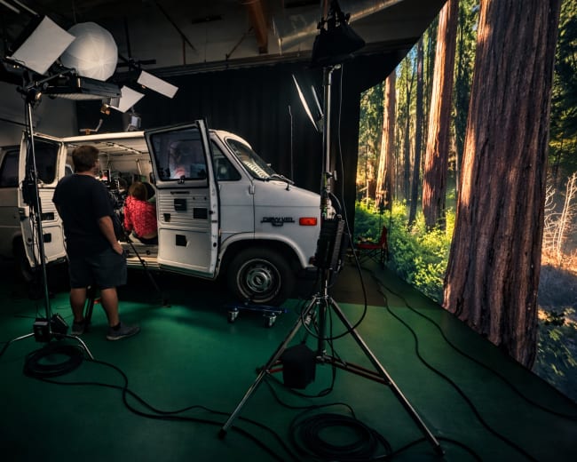 A film crew sets up a scene inside a van, with large lights and cameras, while a backdrop of a forest is displayed behind them.