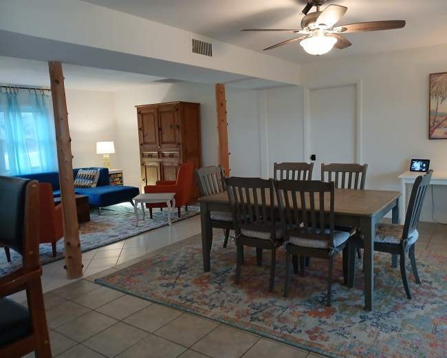 The image shows a dining area with a large table and chairs, surrounded by a living space featuring a couch, a lamp, and a decorative cabinet.