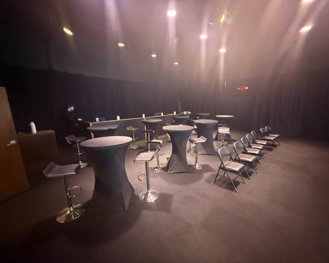 The scene depicts a dimly lit space with several tall tables and chairs arranged in a casual layout against a backdrop of black curtains.