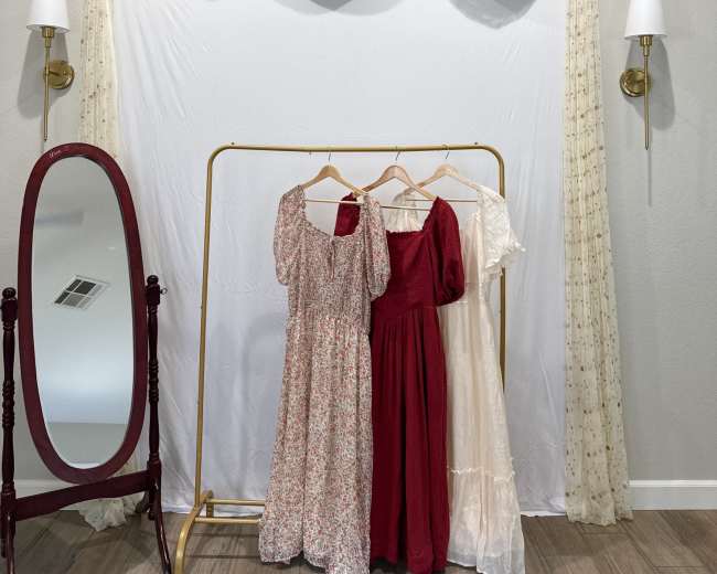 A simple dressing area features a white backdrop with draped fabric, a clothing rack displaying three dresses, and a full-length mirror nearby.
