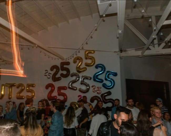 A group of people celebrates in a venue adorned with large glittering "25" balloons.