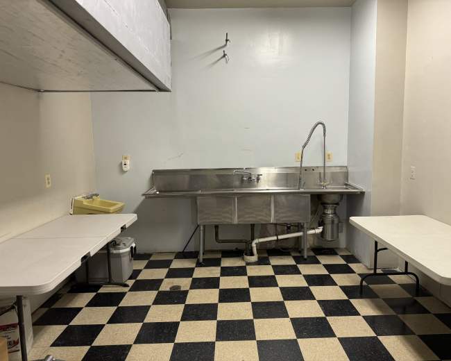 The image shows a small commercial kitchen space with a stainless steel sink, a couple of white tables, and checkered black and beige flooring.